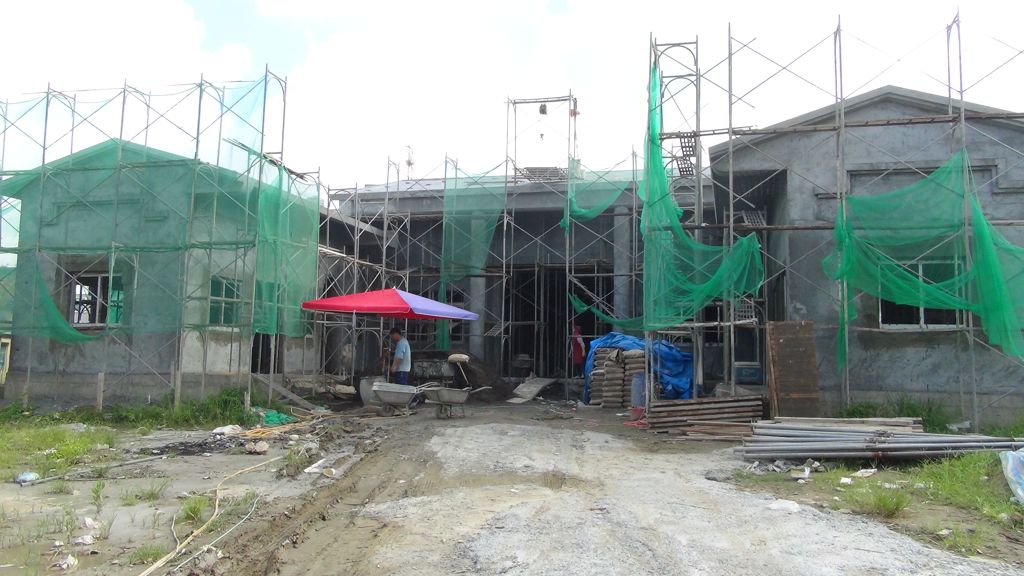 興建中的宗祠正面，滿架鋼管鷹架與綠色安全網，牆體仍是清水混凝土。屋簷下搭著一頂紅藍色帆布傘，傘下兩名工人與兩台獨輪推車，右側堆放水泥袋、藍色帆布與成捆的鷹架鋼管，地面是未整平的泥地。
