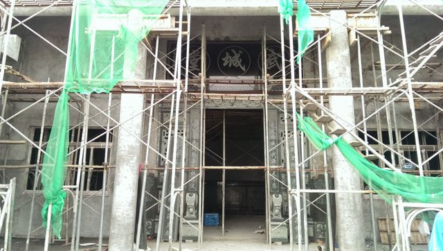 施工中的宗祠正面，滿布鋼管鷹架與綠色安全網。門楣上方已懸「武城堂」堂號匾，門柱兩側的石雕對聯亦已就位，牆面尚為清水混凝土。