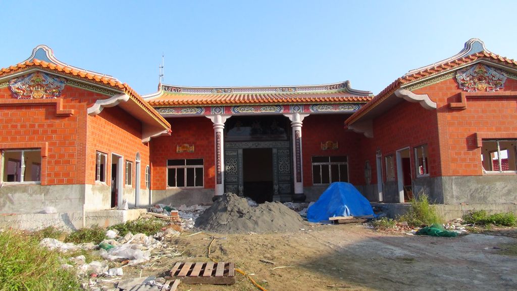 宗祠正面，屋面已完成而內外裝未竟。紅磚牆、橘色琉璃瓦屋脊與兩側山牆交趾陶皆已就位，中央門廊立柱與雕花大門亦已裝上；門前堆著一堆灰砂與藍色帆布，兩側窗洞尚未裝窗，地面散落建材與棧板。