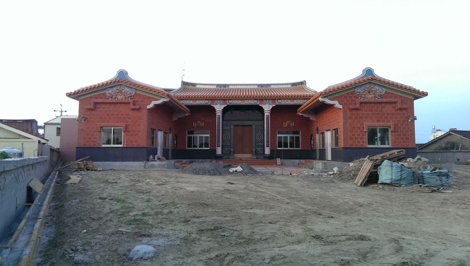 黃昏時的武城堂宗祠正面。天色轉為淺灰，紅磚牆與橘瓦屋脊已完成，中央大門已裝上木門，門前庭院整平為土坪；左側圍牆施工中，右側堆放木料與覆蓋綠色帆布的材料。